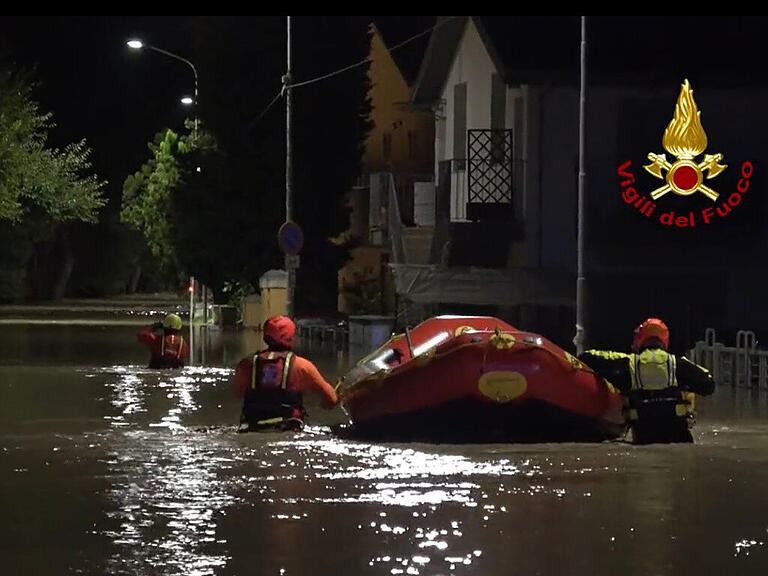 HANDOUT - Feuerwehrleute schieben ein Schlauchboot durch eine überflutete Straße, um Menschen aus den Häusern zu retten. Foto: -/Vigili del Fuoco/dpa - ACHTUNG: Nur zur redaktionellen Verwendung im Zusammenhang mit der aktuellen Berichterstattung und nur mit vollständiger Nennung des vorstehenden Credits